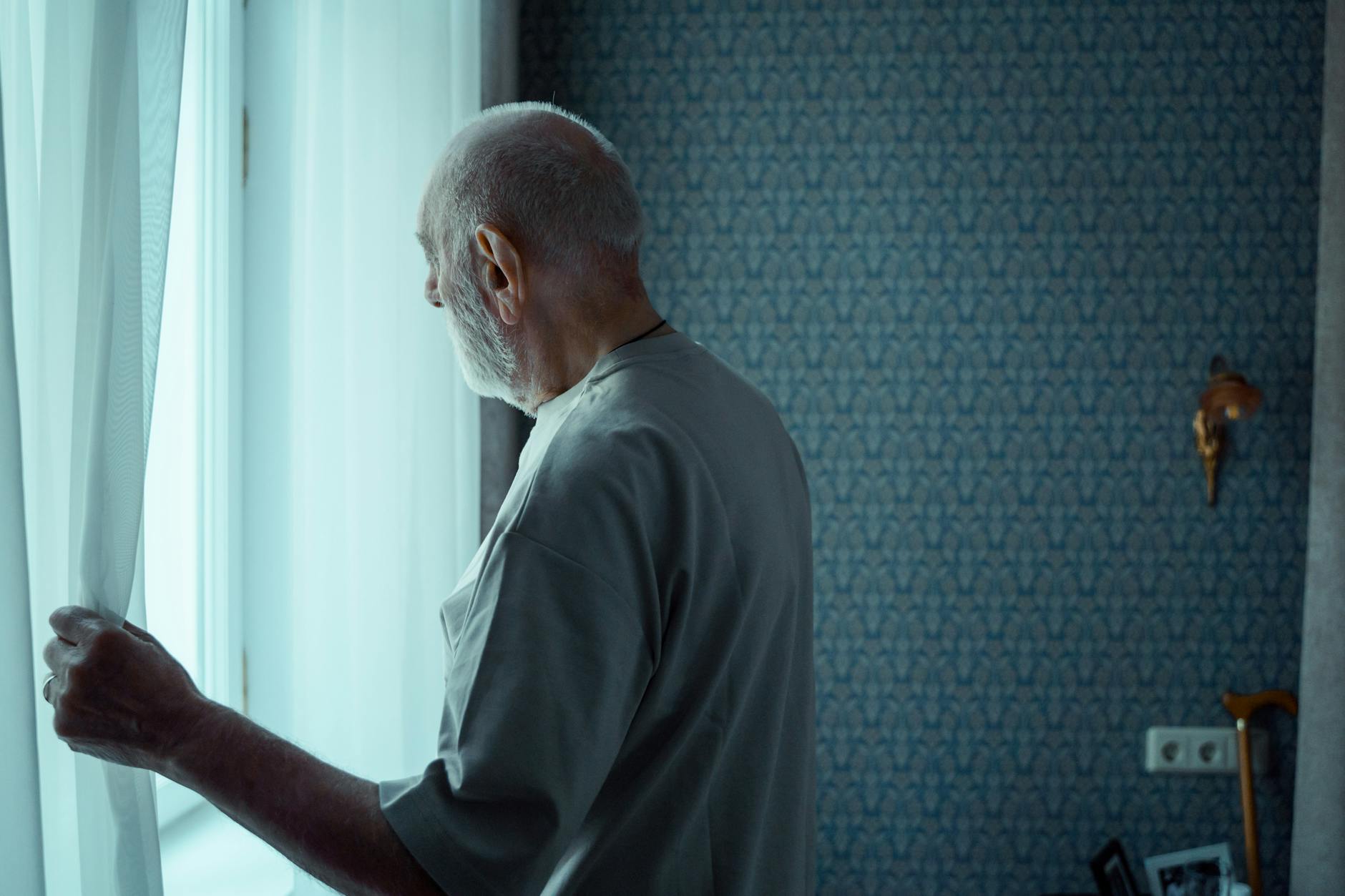 Social Isolation and Untreated Hearing Loss Senior man standing alone looking out a window, reflecting the social isolation that untreated hearing loss can cause